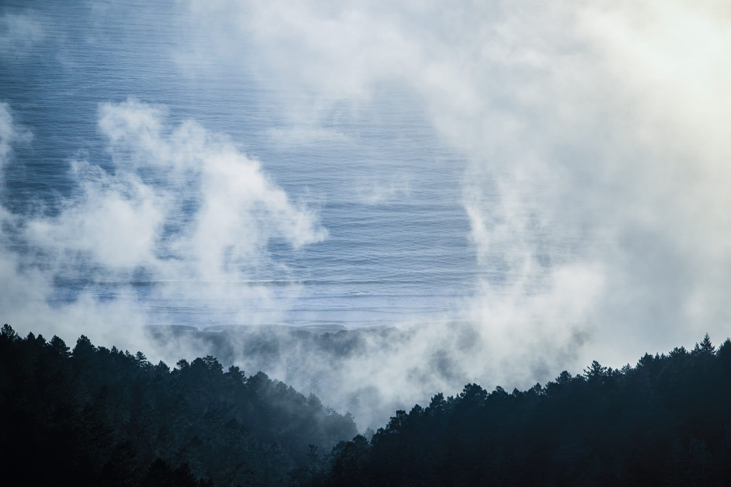 Fog drifting over the ocean above a dark forested ridge, fine art photograph by Jason Bowman