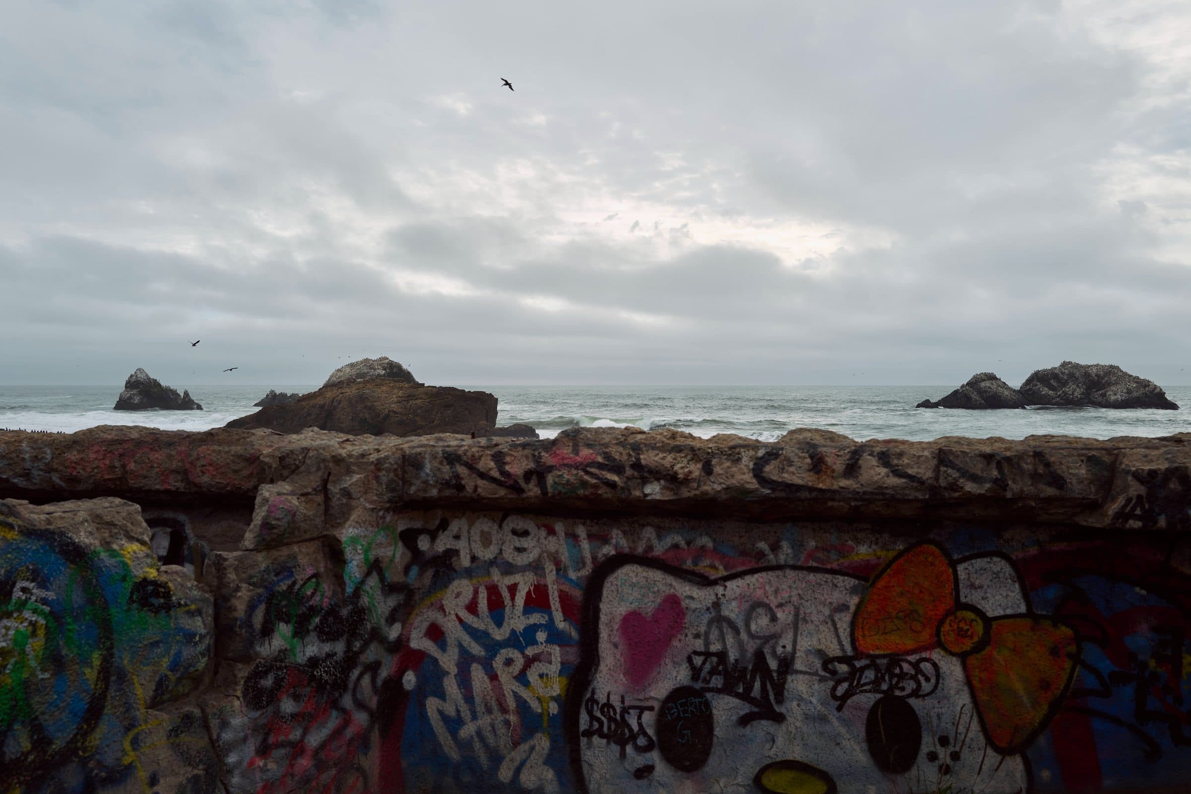 Graffiti-covered stone wall facing sea rocks under an overcast sky, fine art photograph by Jason Bowman