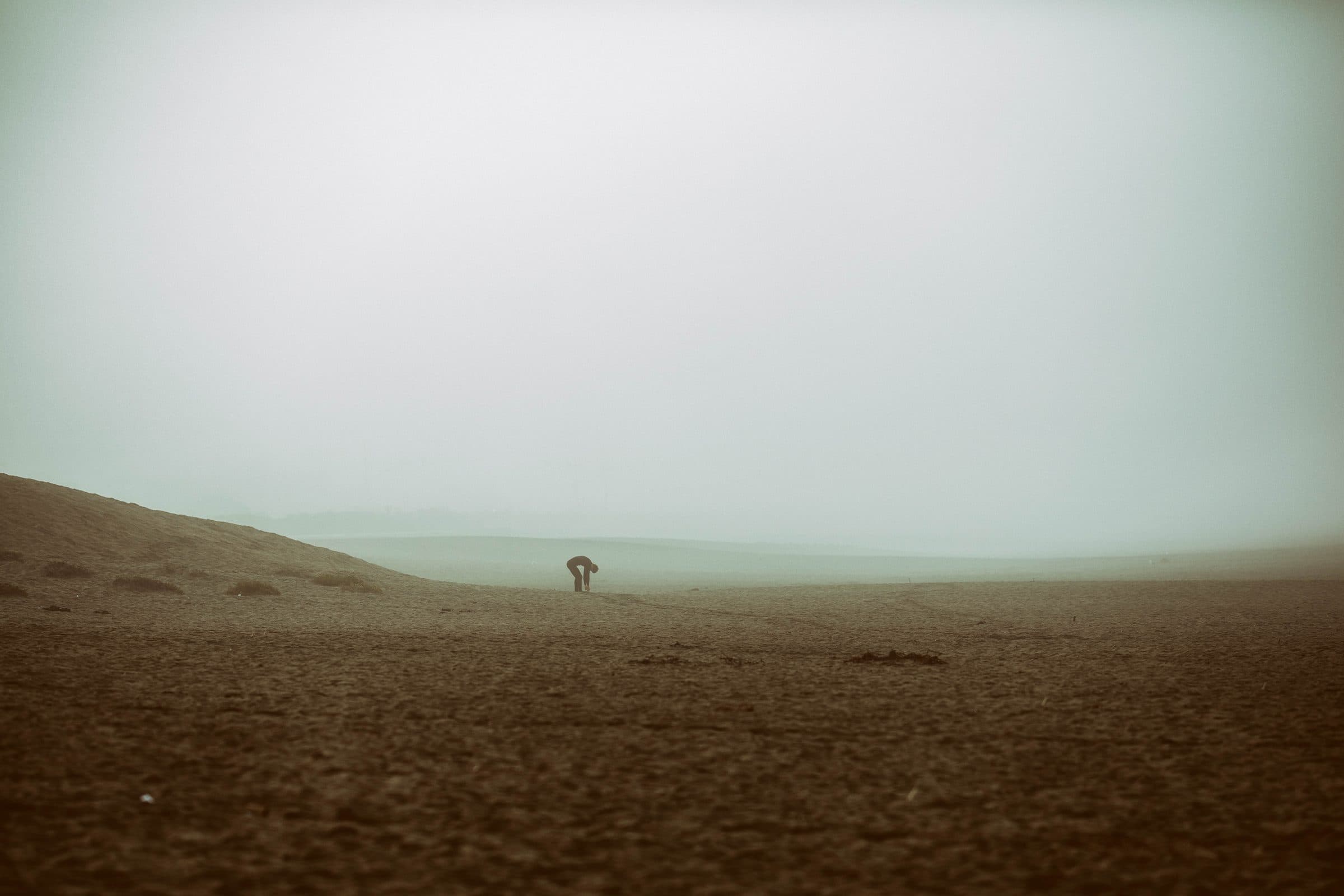 A lone figure bending down on a fog-covered beach, fine art photograph by Jason Bowman