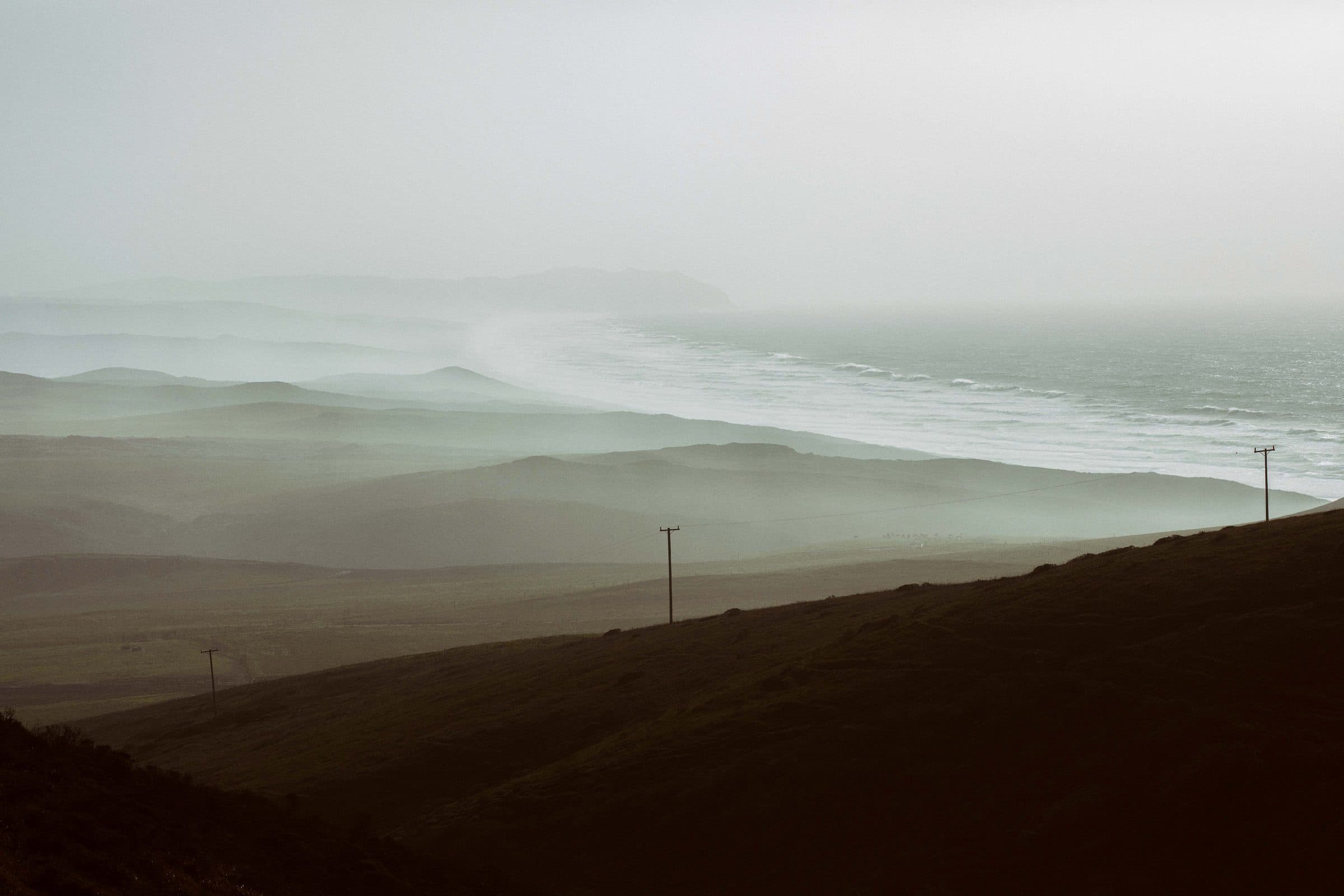Misty layered hills and coastline with telephone poles on a slope, fine art photograph by Jason Bowman