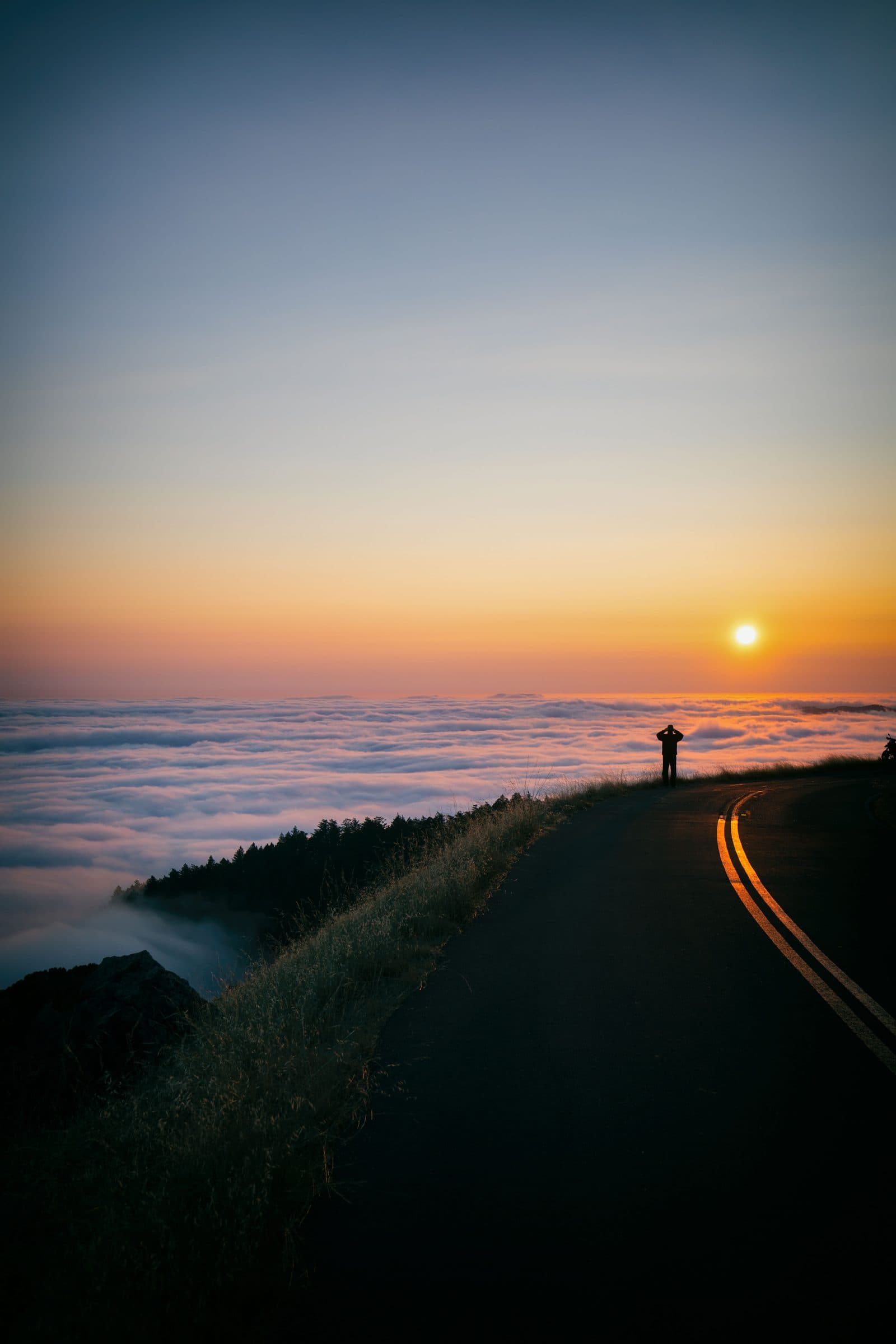 A figure silhouetted at sunset on a ridge road above a sea of fog, fine art photograph by Jason Bowman