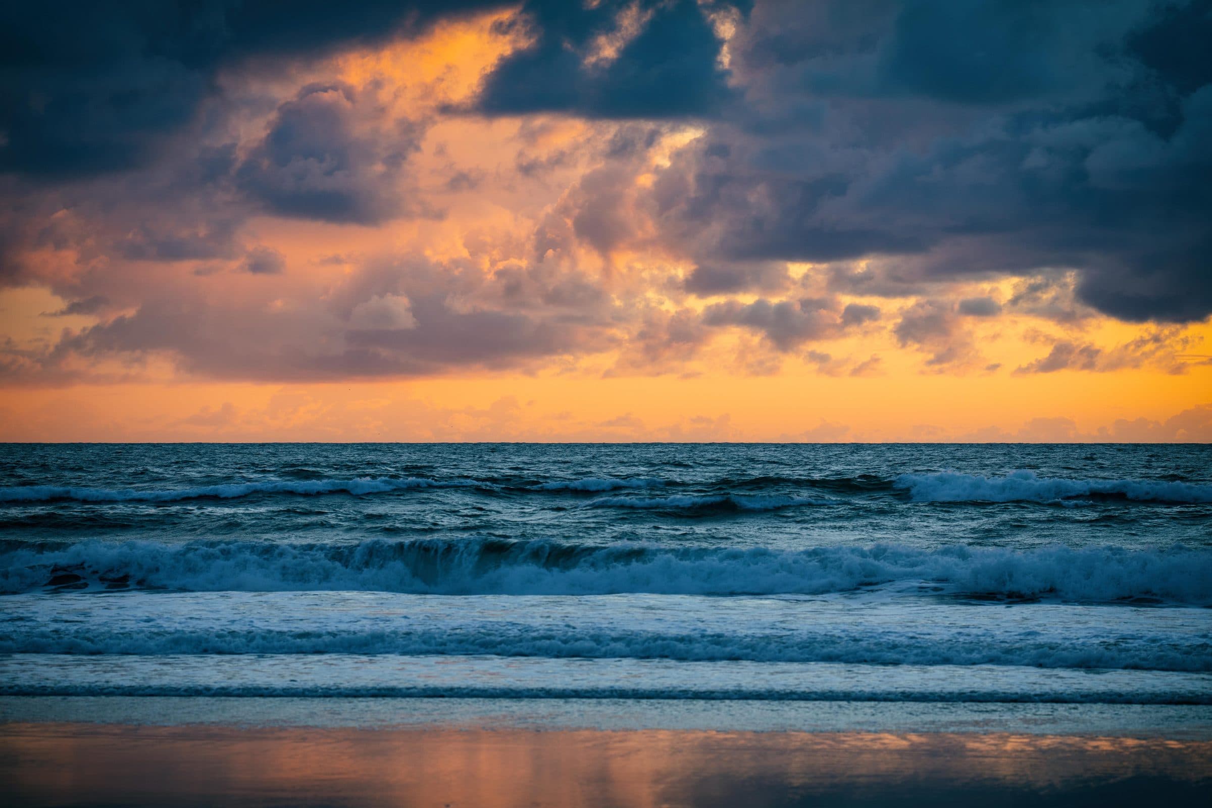 Orange sunset through storm clouds over breaking waves, fine art photograph by Jason Bowman